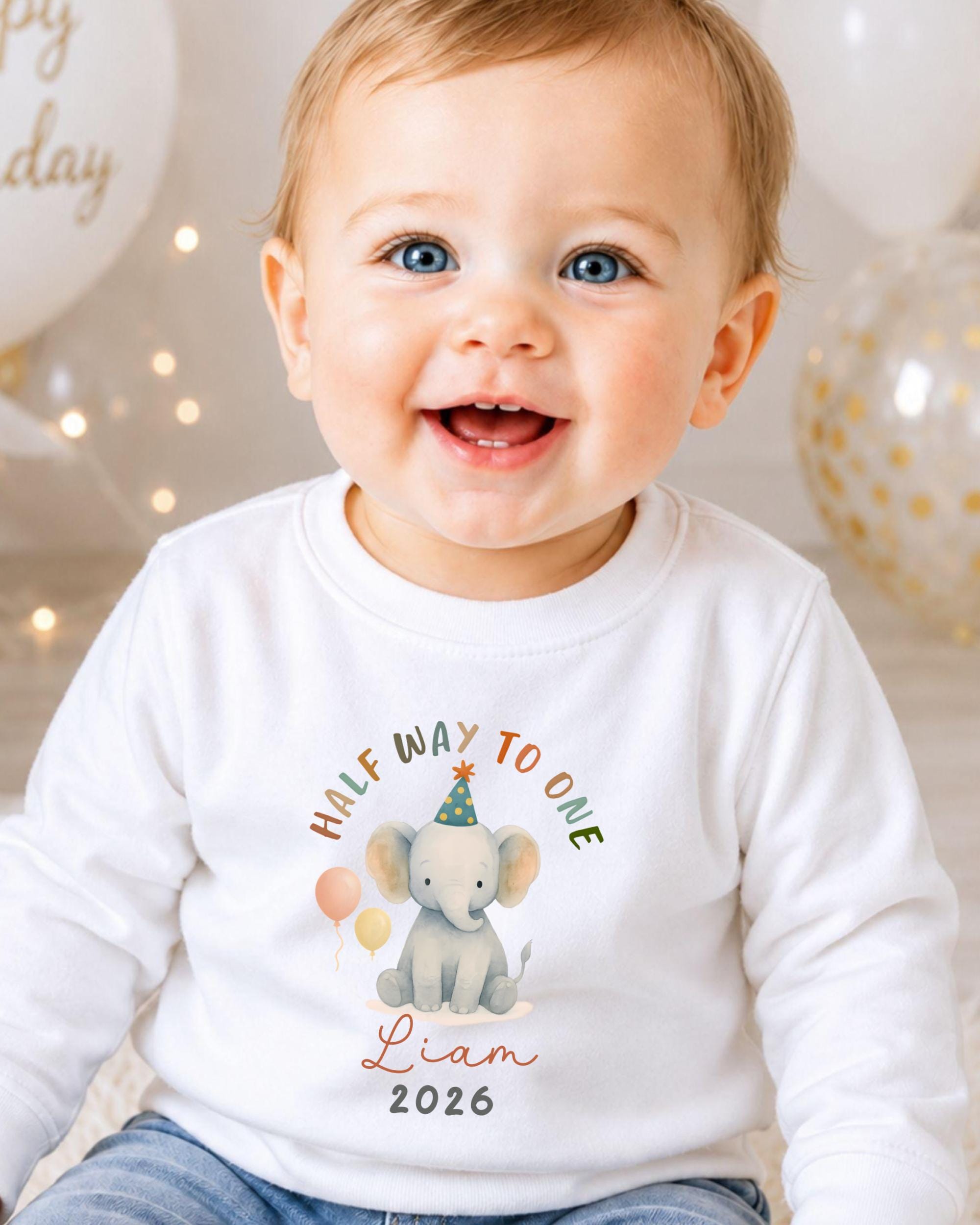 A smiling baby boy wearing a white shirt with a graphic of an elephant and the text "Half way to one Liam 2026" printed on it.