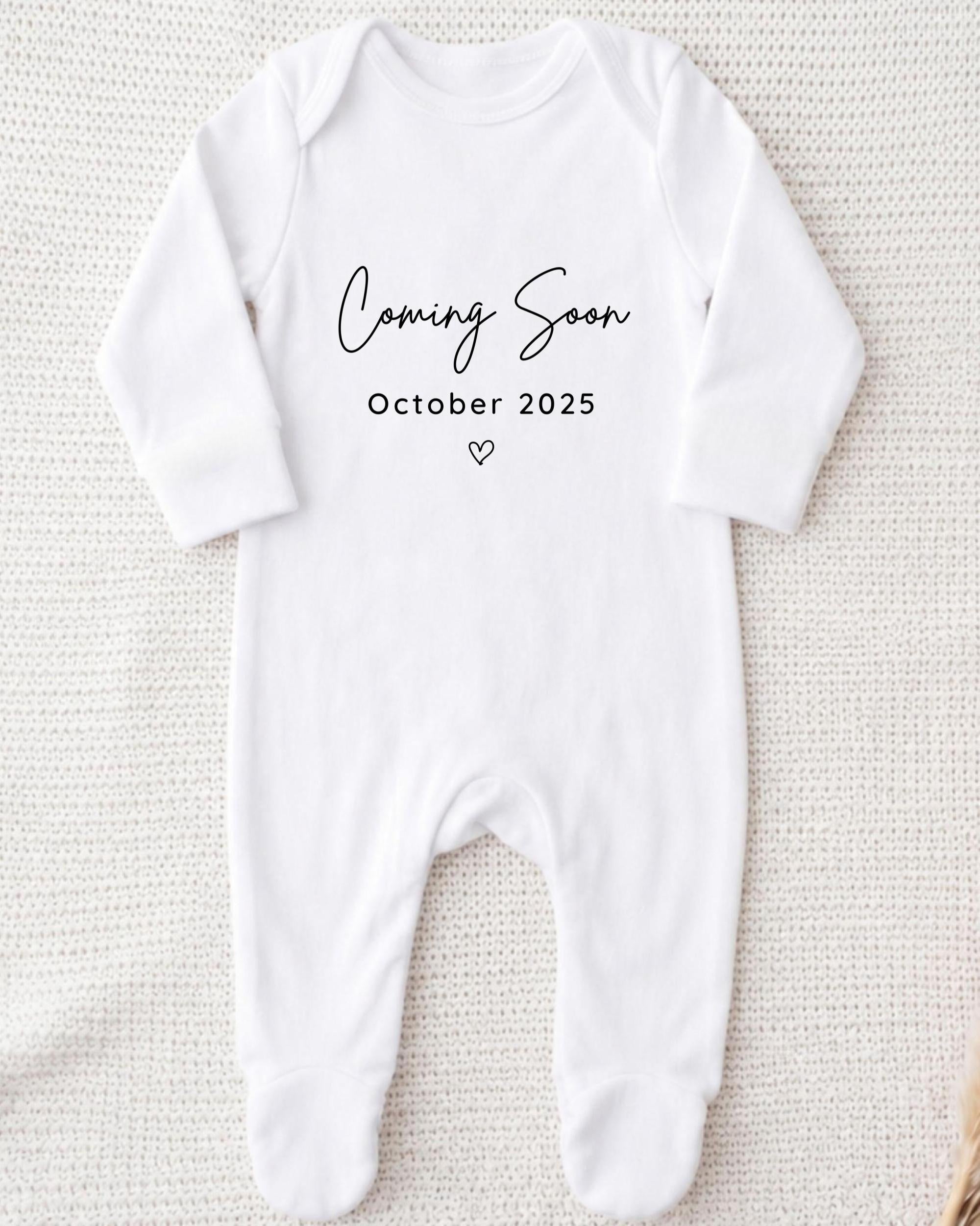 A white onesie with the text "Coming Soon October 2025" printed on it.