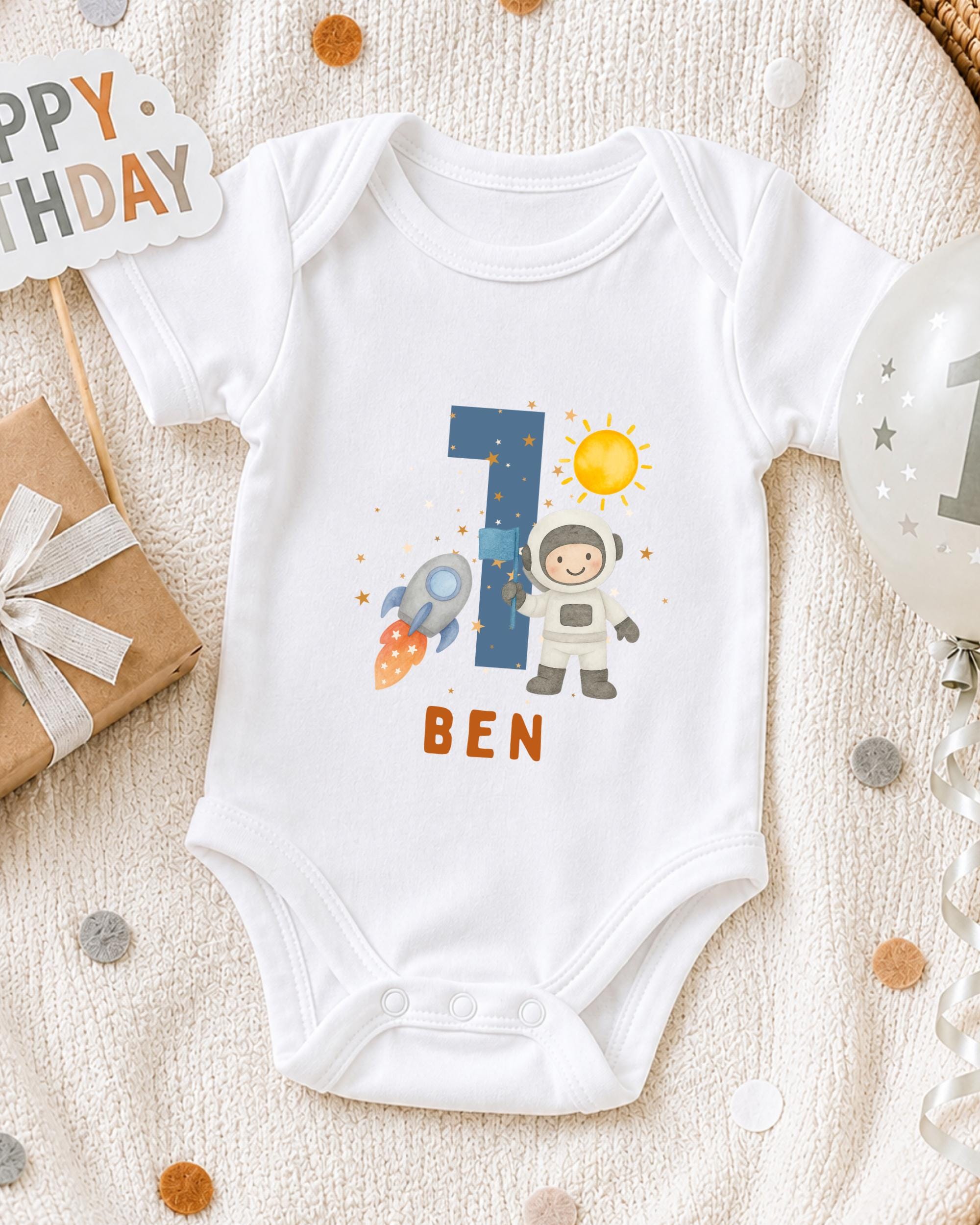 A white onesie with a space theme design, featuring a rocket ship, a person in a spacesuit, and the number 1. The word "BEN" is written below the image.