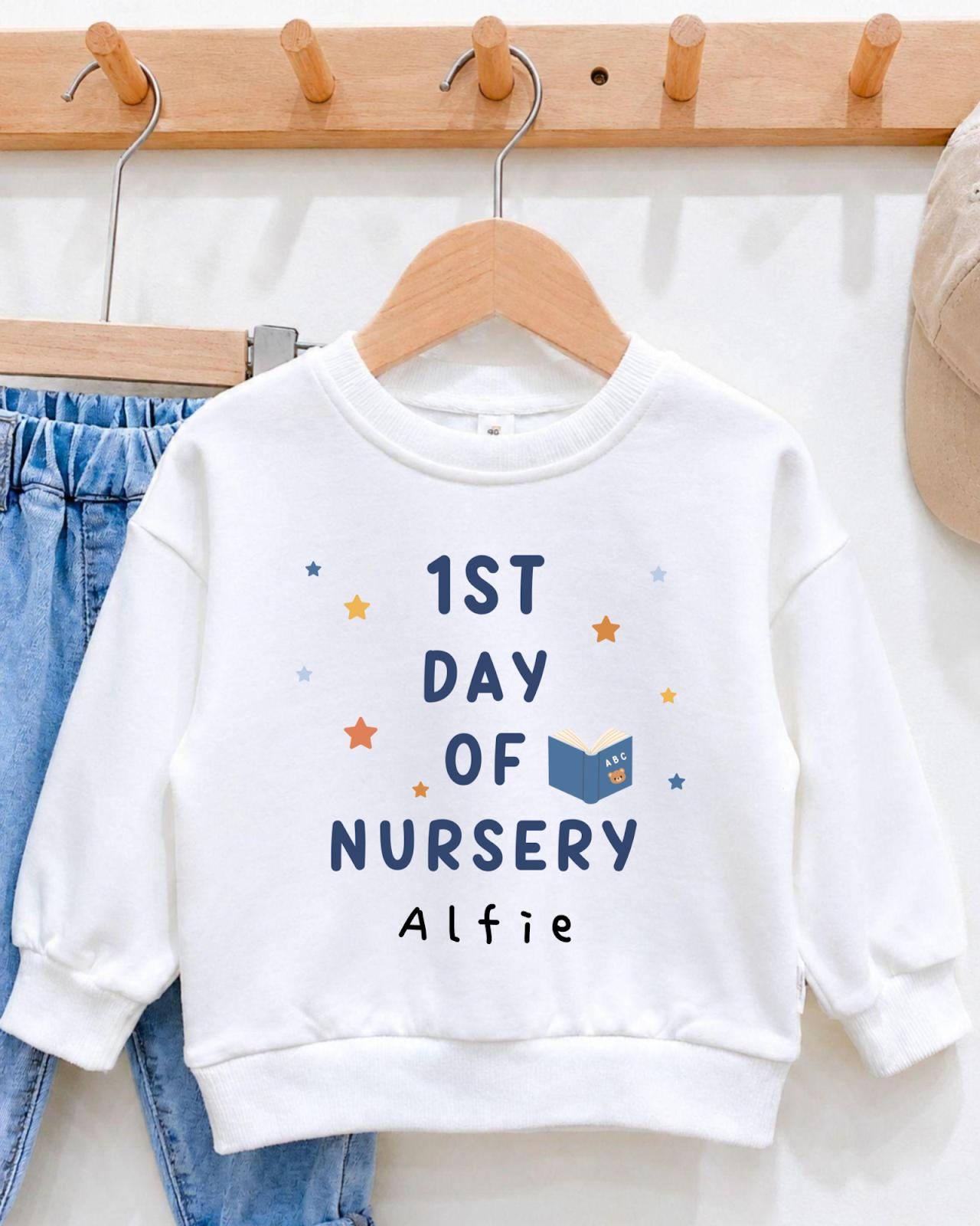A white sweatshirt with the text "1st DAY OF NURSERY" printed on it, along with a small book icon and stars. The sweatshirt is hanging on a wooden hanger.