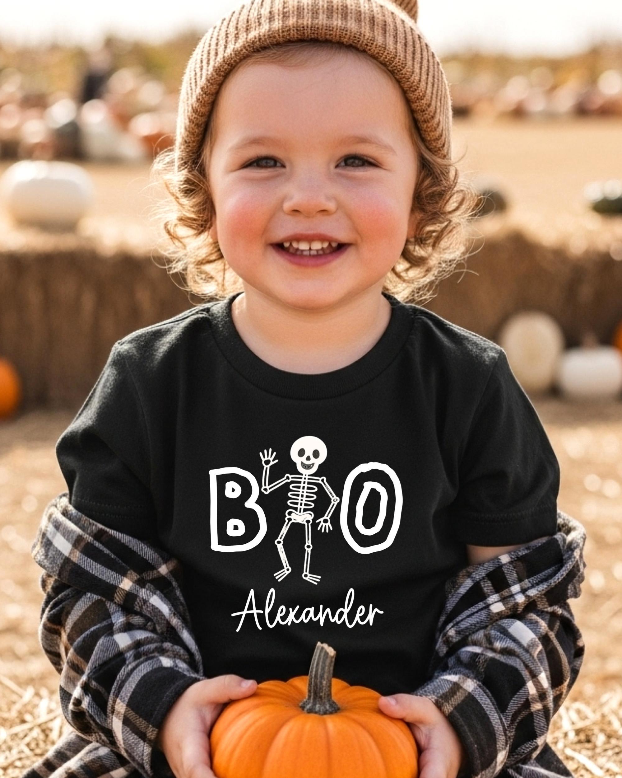 A young child wearing a black t-shirt with a skeleton design and the name "Alexander" printed on it, holding an orange pumpkin.