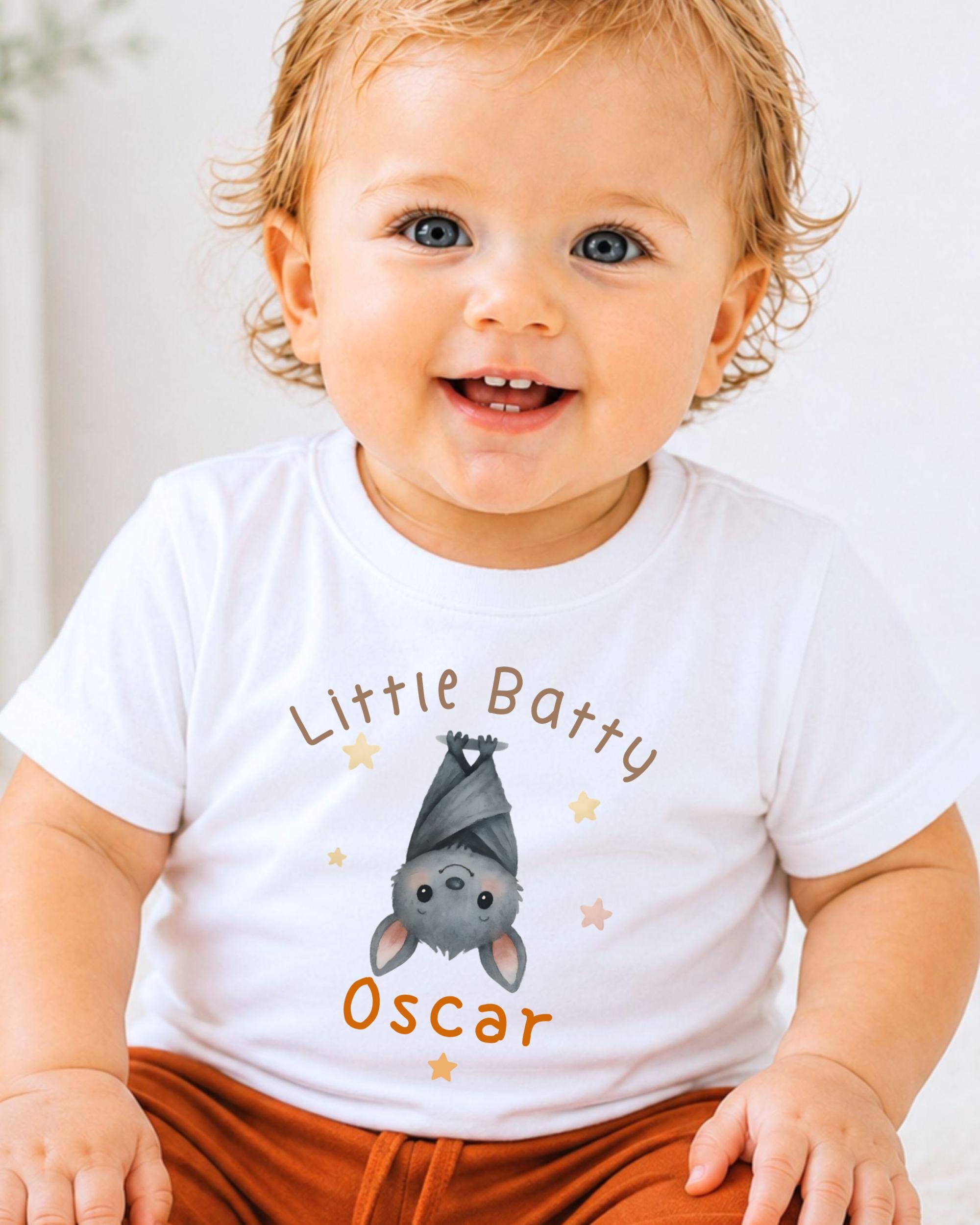 A smiling baby wearing a white t-shirt with a bat graphic and the name "Oscar" printed on it.