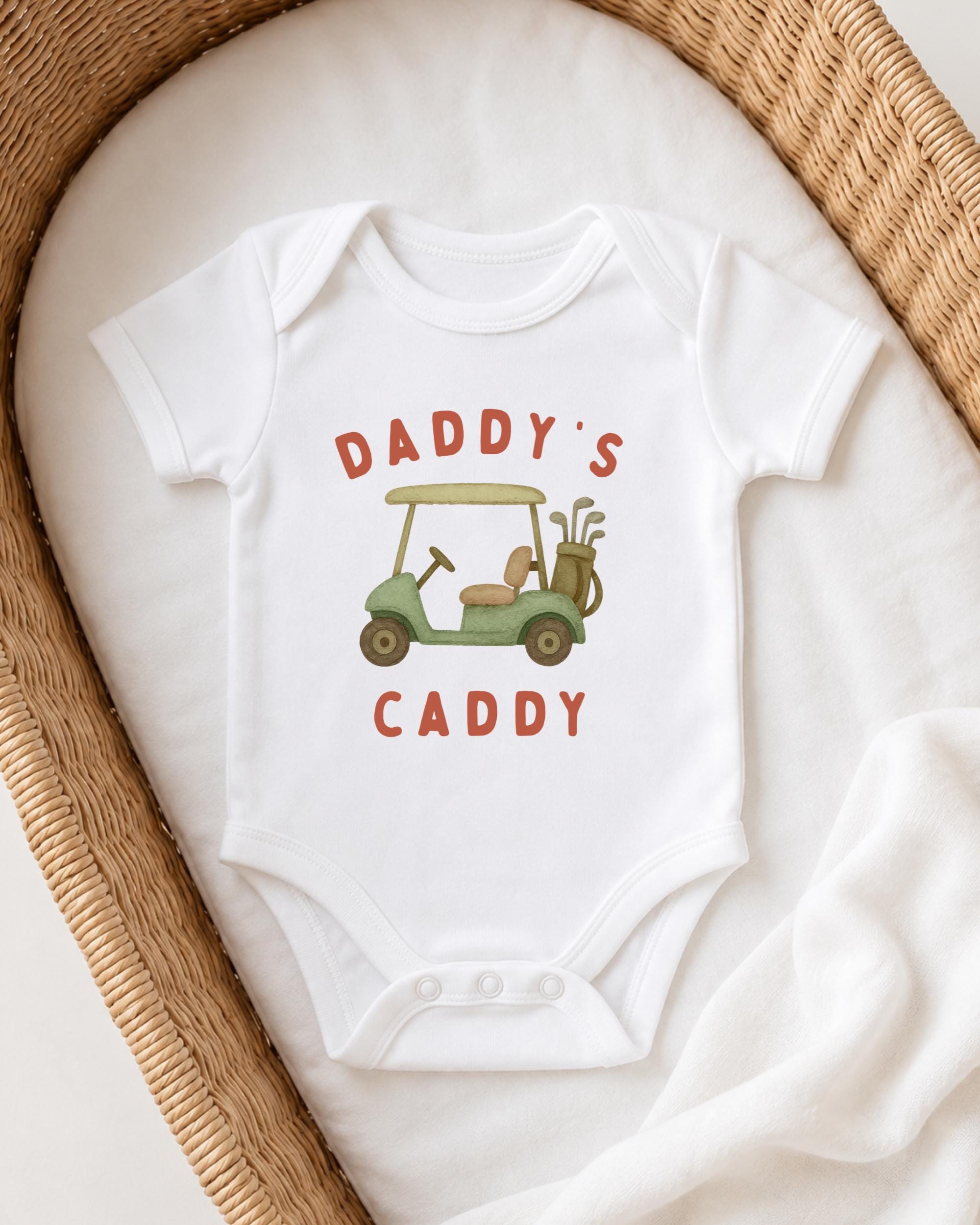 A white onesie with a golf cart design and the text "DADDY'S CADDY" printed on it, laid out on a white cloth.