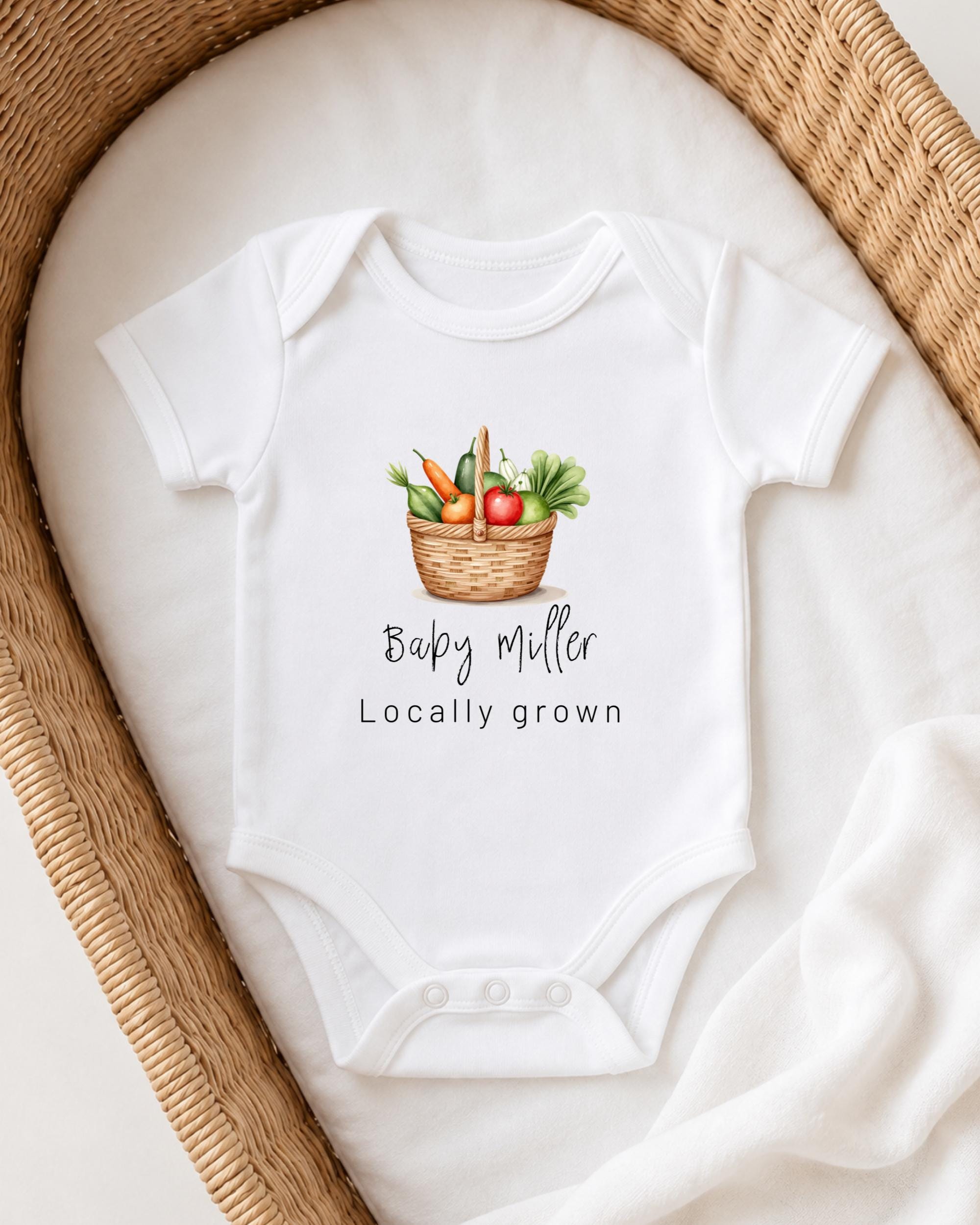 A white baby onesie with a basket of vegetables printed on it, placed on a white blanket.