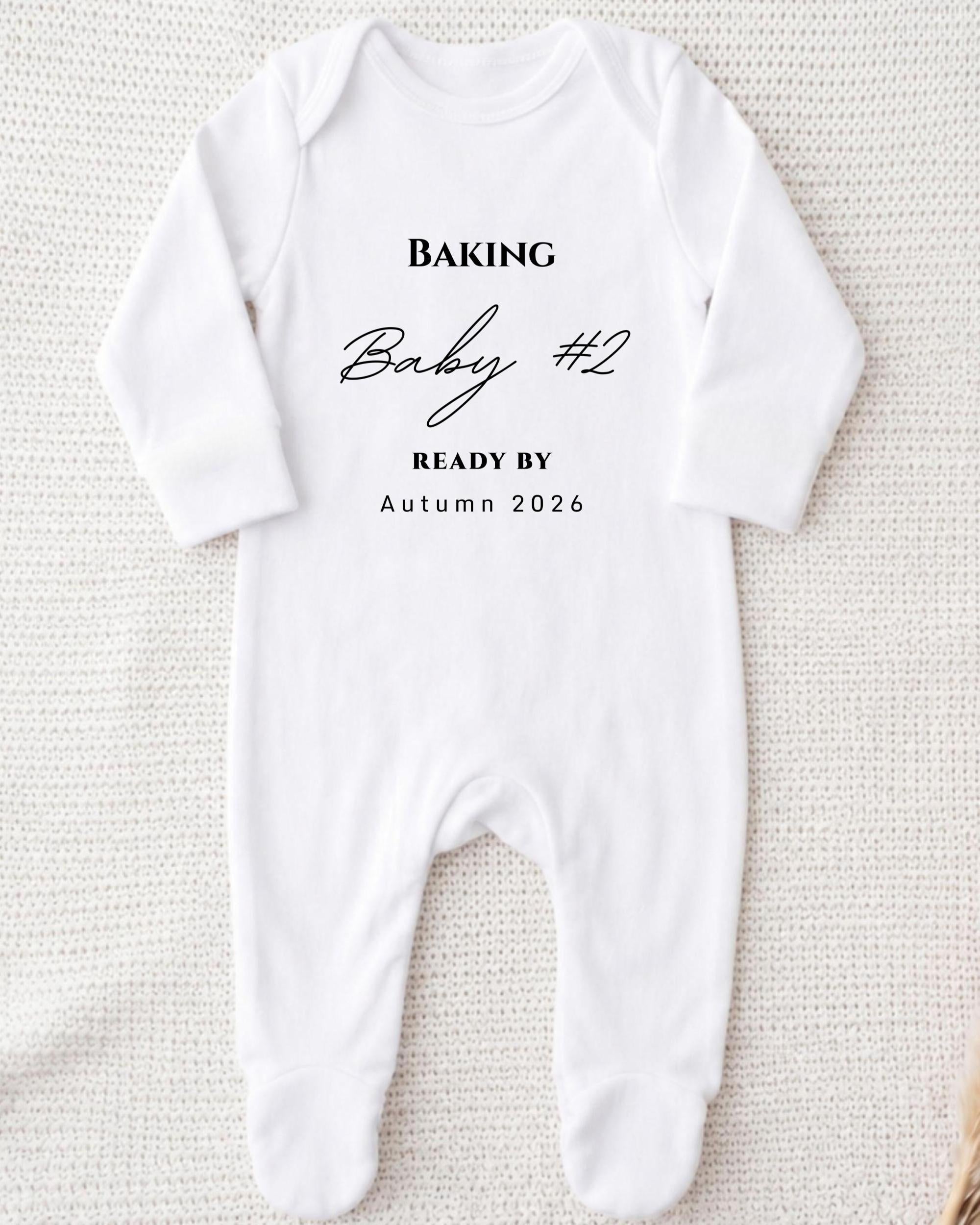 A white onesie with the text "BAKING Baby #2 READY BY Autumn 2026" printed on it.