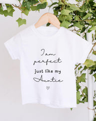 A white t-shirt with black text that reads "I am perfect just like my Auntie" hanging on a wooden hanger, with green ivy leaves in the background.