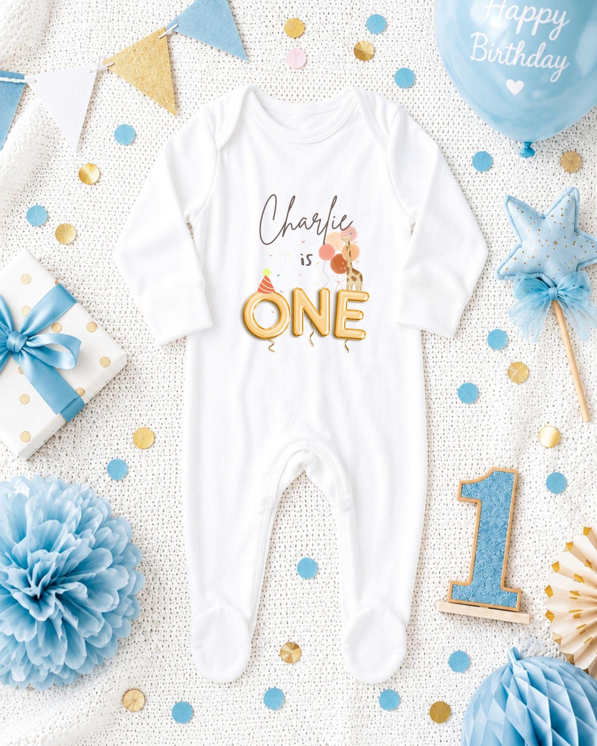 A white onesie with the text "Charlie is one" printed on it, surrounded by various baby-themed items such as a blue balloon, a star, confetti, and a gift box.