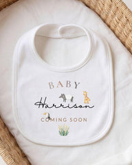 A white baby bib with the text "BABY Harrison" and "COMING SOON" printed on it, along with illustrations of a giraffe and a horse.