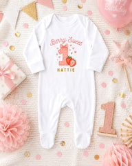 A white onesie with a berry and a number one design, surrounded by pink and gold decorations, confetti, and a pink balloon with the text "Happy Birthday".