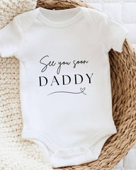 A white baby onesie with the text "See you soon Daddy" printed on it, placed on a woven basket.