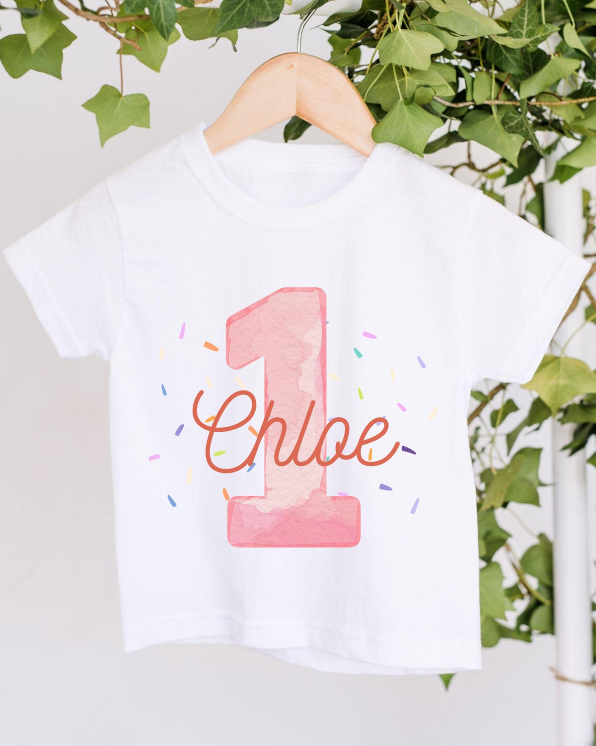 A white t-shirt with a pink number one and the name "Chloe" printed on it, hanging on a wooden hanger.
