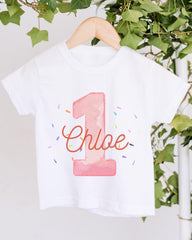 A white t-shirt with a pink number one and the name "Chloe" printed on it, hanging on a wooden hanger.
