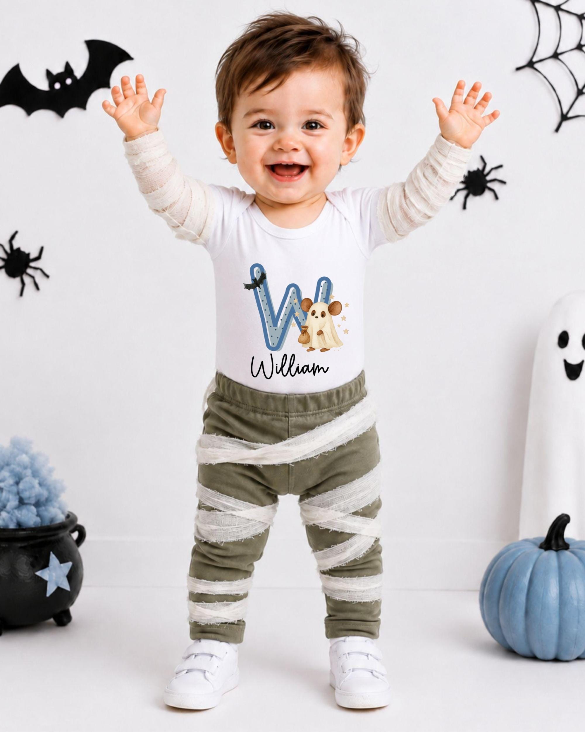A smiling baby boy wearing a white shirt with a letter "W" and striped pants, standing in front of Halloween decorations.