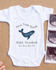 A white baby onesie with a graphic of a blue whale and the text "Sea You Soon Baby Seahell Due December 2027" printed on it, along with a photograph of an ultrasound image of a baby.
