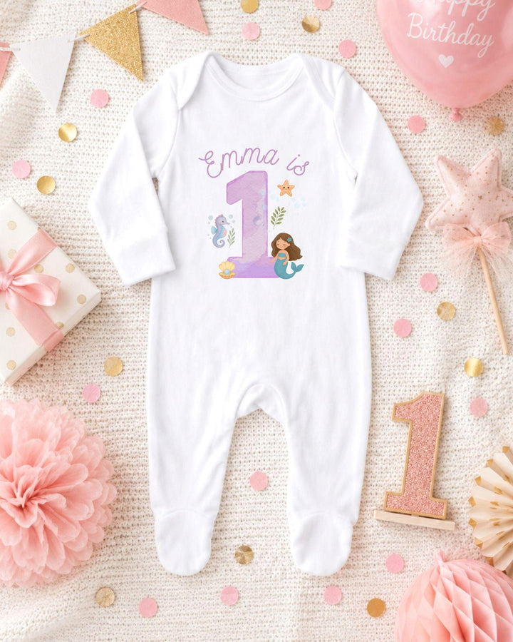 A white bodysuit with a mermaid design and the number 1, surrounded by pink and gold decorations, including a pink star, confetti, and a pink ribbon.