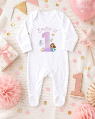 A white onesie with a mermaid design and the number 1, surrounded by pink and gold decorations, including a pink star, confetti, and a pink ribbon.