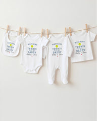 A row of four white baby clothes with the text "WATCHING TENNIS DADDY Daddy & Ava" printed on them, hanging on a clothesline.