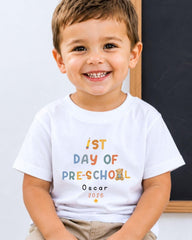 A young boy wearing a white t-shirt with text that says "1st DAY OF PRE-SCHOOL Oscar 2006" smiling at the camera.
