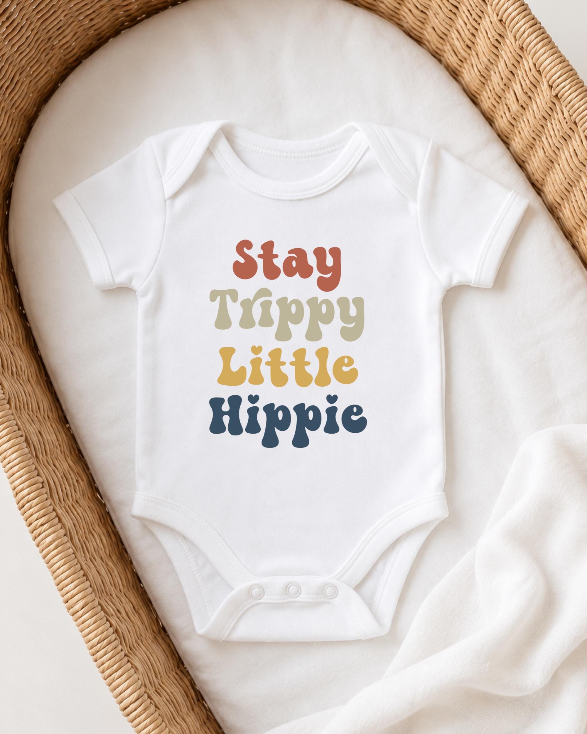 A white baby onesie with the text "Stay Trippy Little Hippie" printed on it, laid out on a white blanket in a wicker basket.