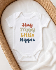 A white baby onesie with the text "Stay Trippy Little Hippie" printed on it, laid out on a white blanket in a wicker basket.