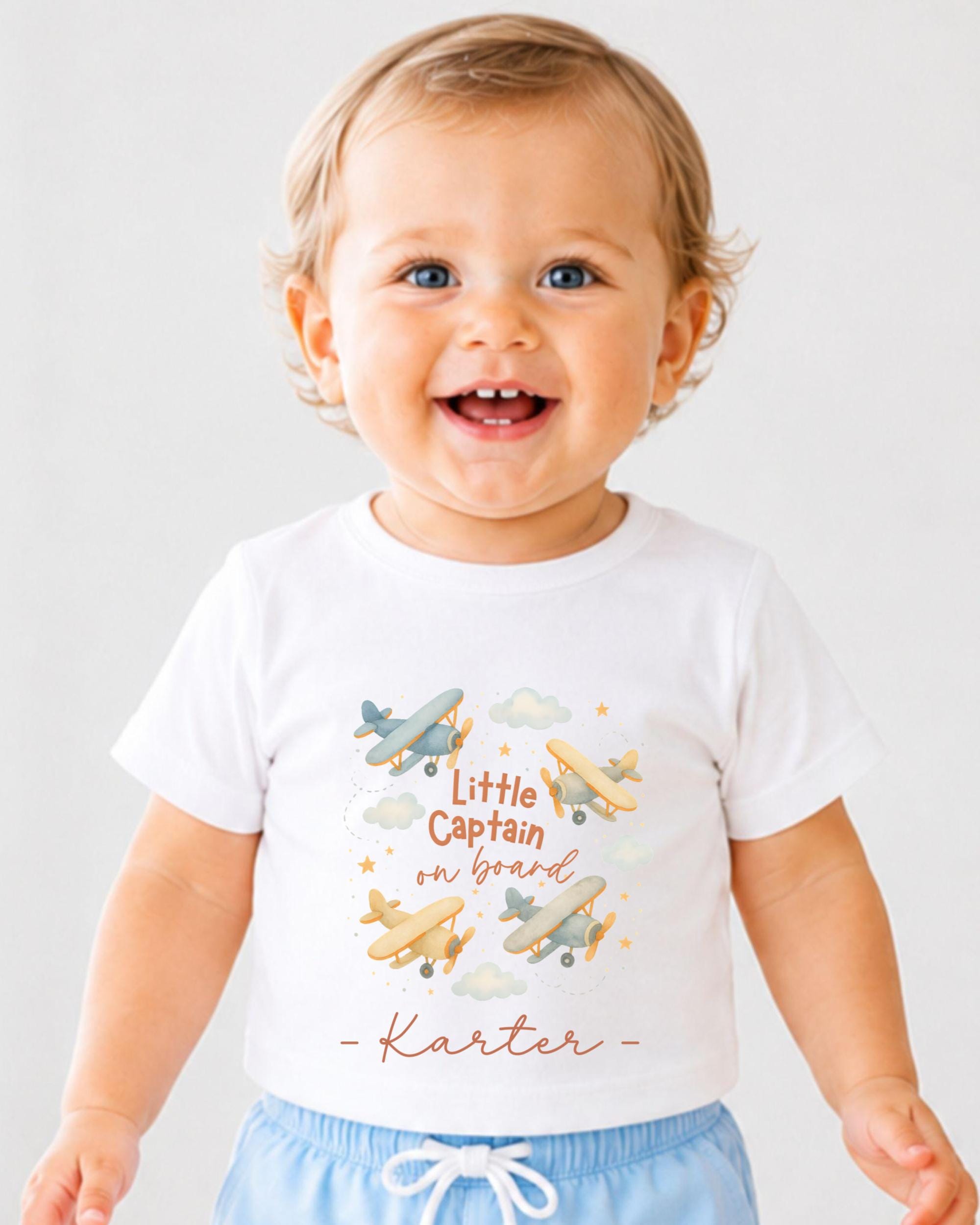 A smiling baby boy wearing a white t-shirt with a graphic design of airplanes and the text "Little Captain on board" printed on it.