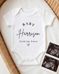 A white baby onesie with the text "BABY Harrison Coming Soon" printed on it, along with a heart symbol. The onesie is placed on a white surface, and there are two black and white images of an ultrasound image next to it.