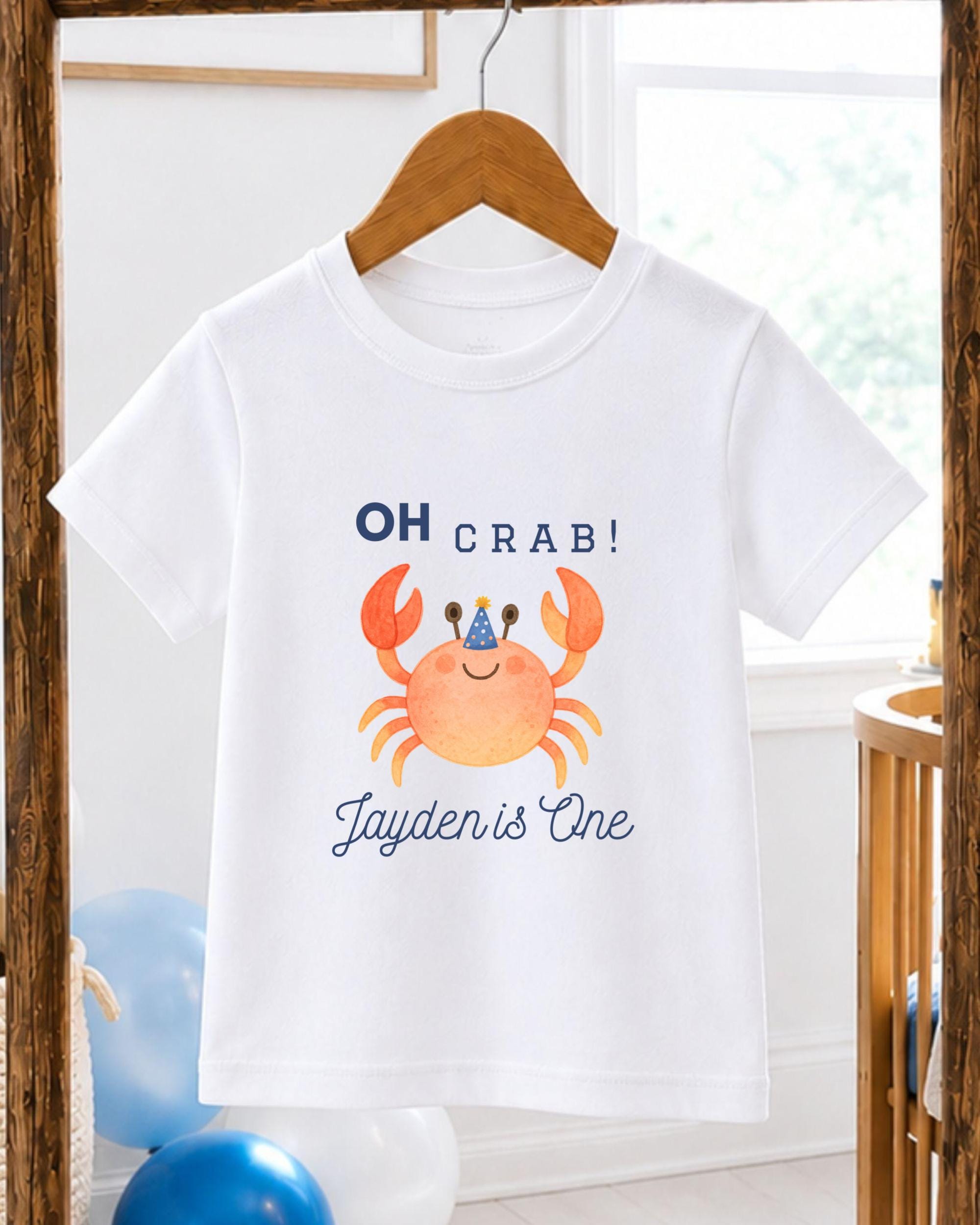 A white t-shirt with a crab design and text printed on it, hanging on a wooden hanger.
