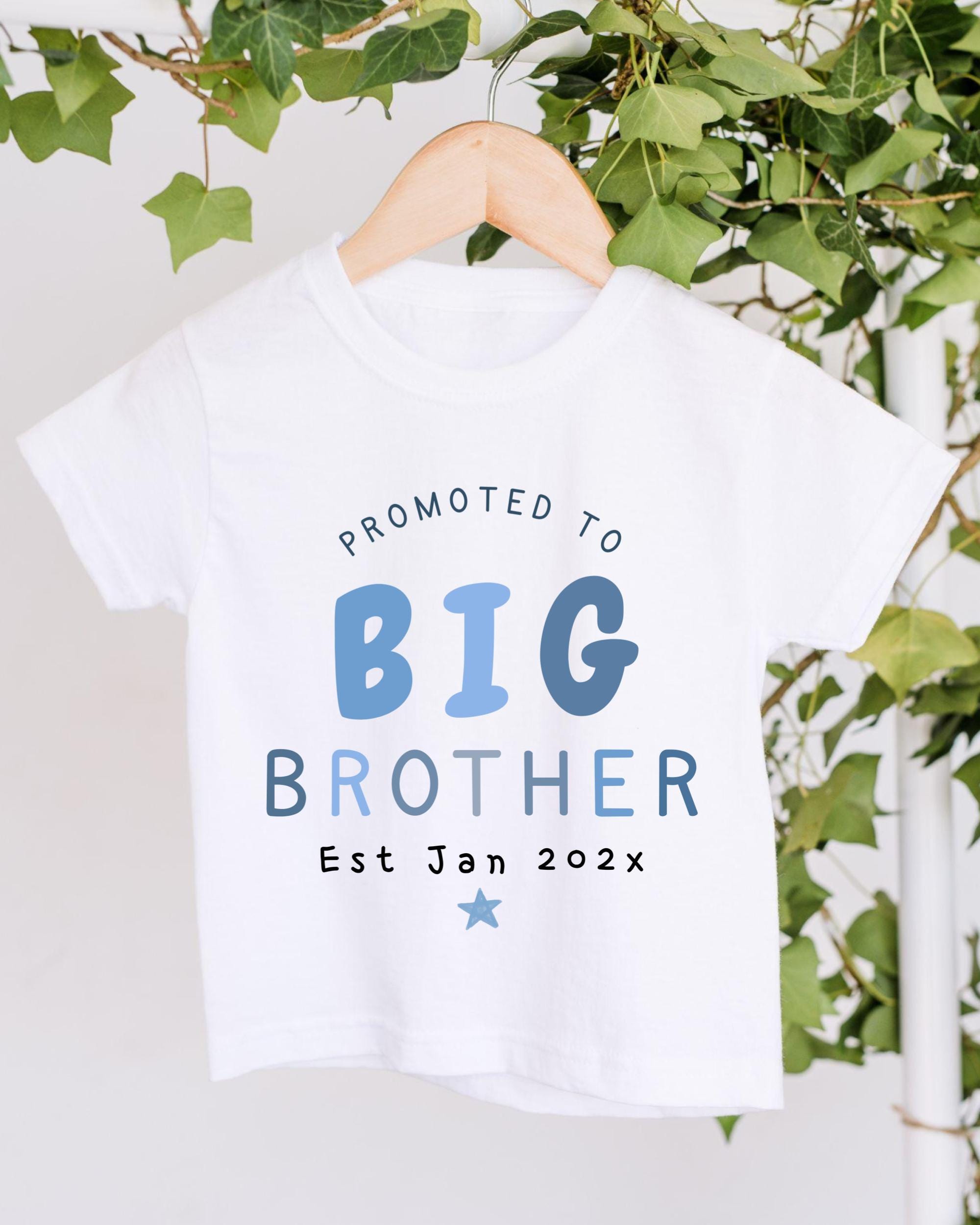 A white t-shirt with text that reads "PROMOTED TO BIG BROTHER Est Jan 202x" hanging on a wooden hanger.