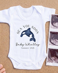 A white onesie with a graphic of a whale and the text "SEA YOU SOON Baby Whalley Summer 2026" printed on it, along with a photograph of an ultrasound image of a baby whale.