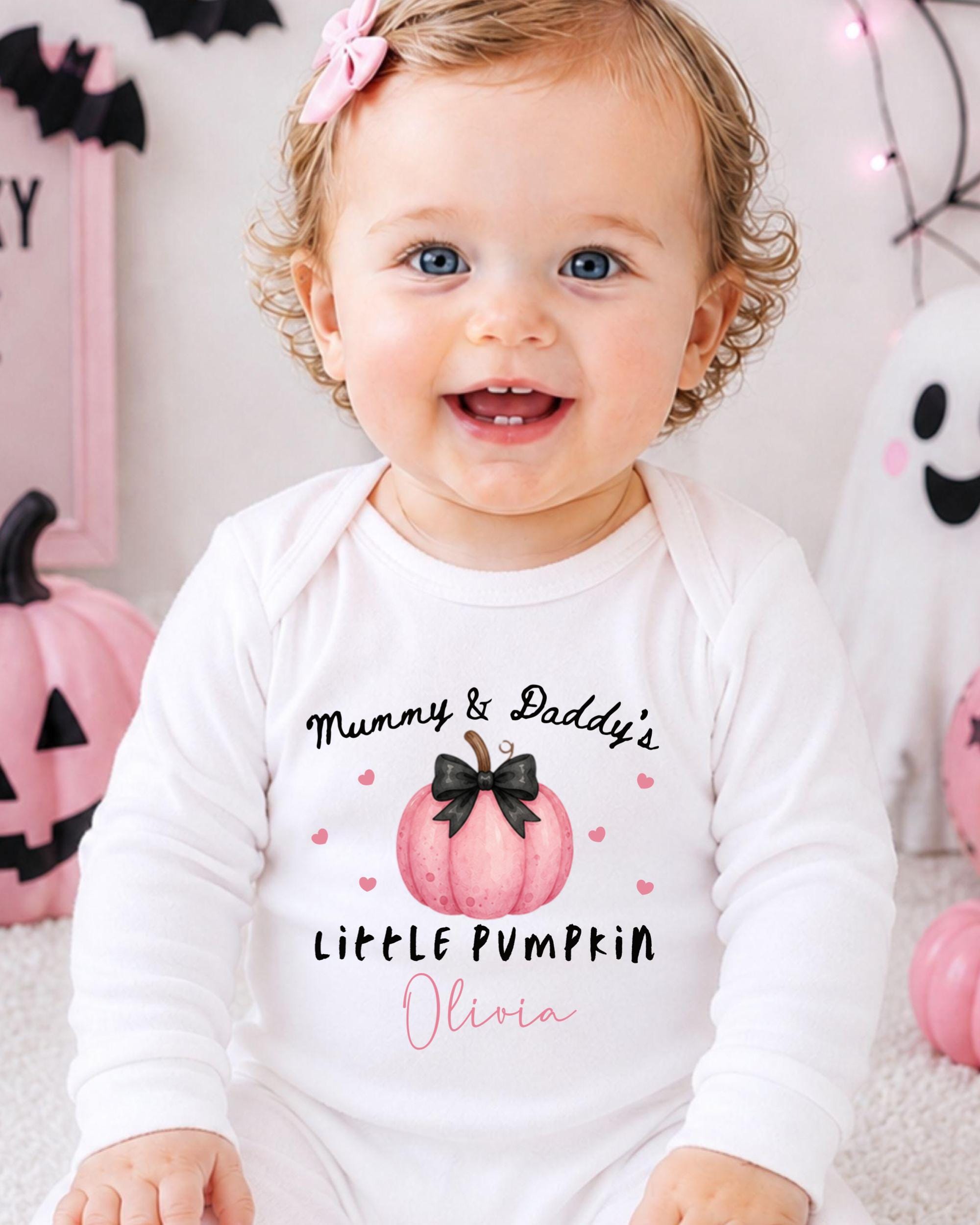 A smiling baby girl wearing a white onesie with a pumpkin design and text that says "Mummy & Daddy's Little Pumpkin Olivia".