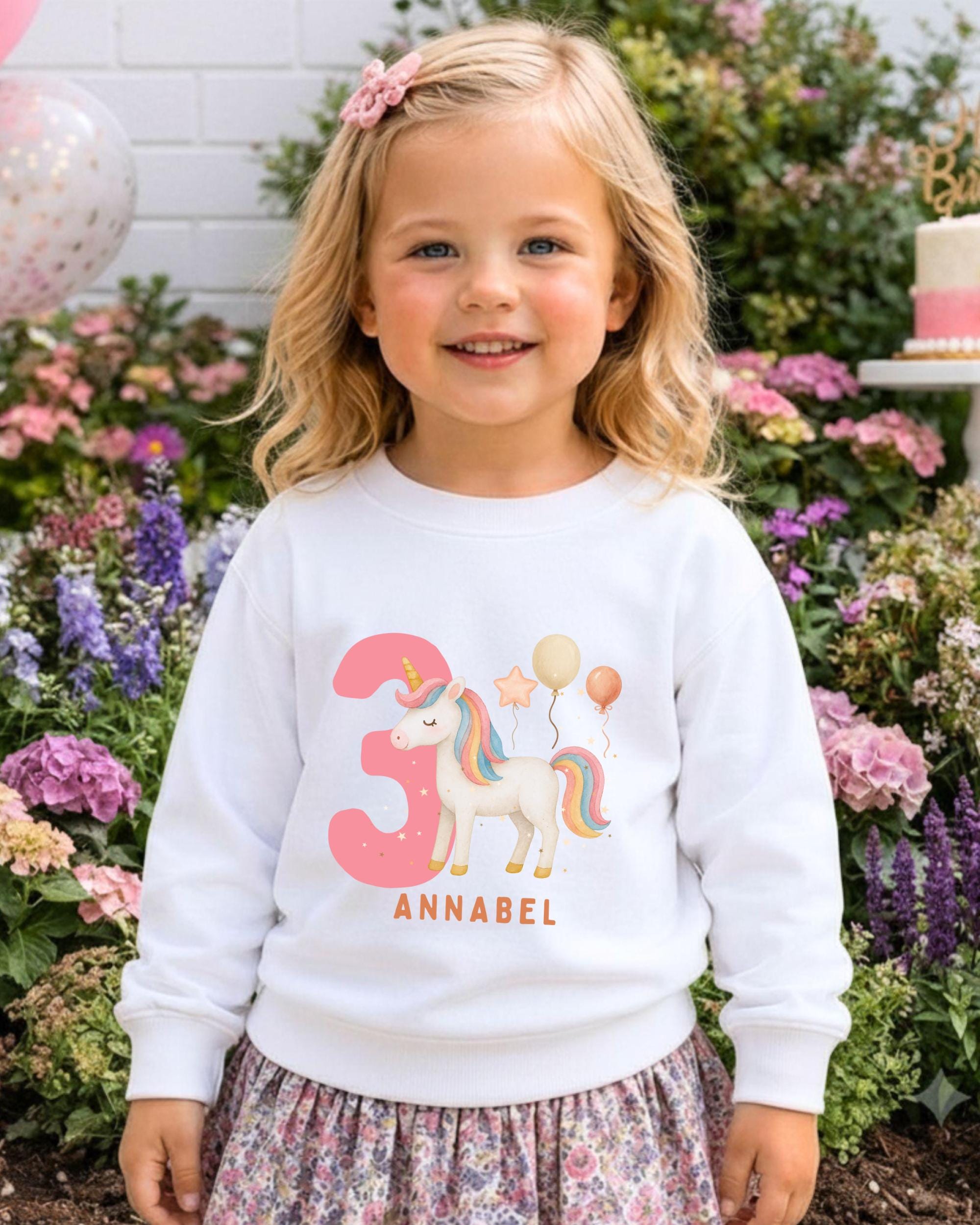 A young girl wearing a white sweater with a unicorn and number 3 printed on it, standing in front of a garden with colorful flowers.
