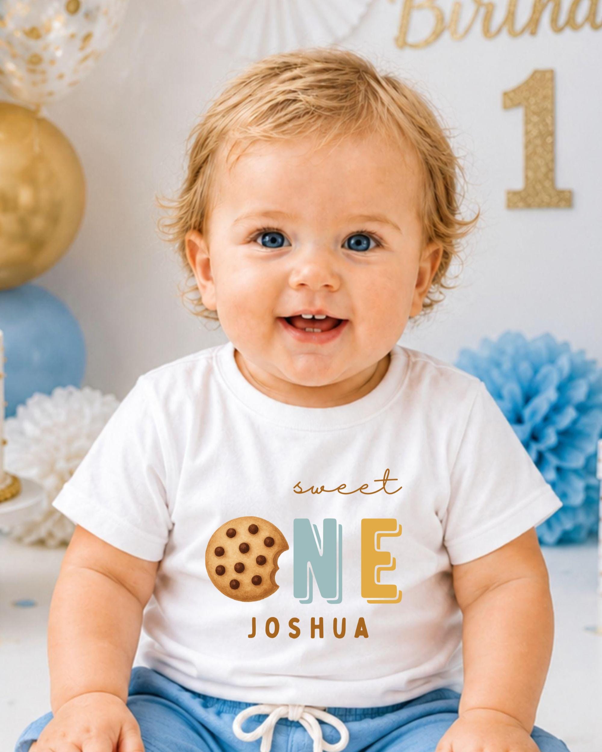 A smiling baby boy wearing a white t-shirt with a cookie and the text "Sweet One Joshua" printed on it.