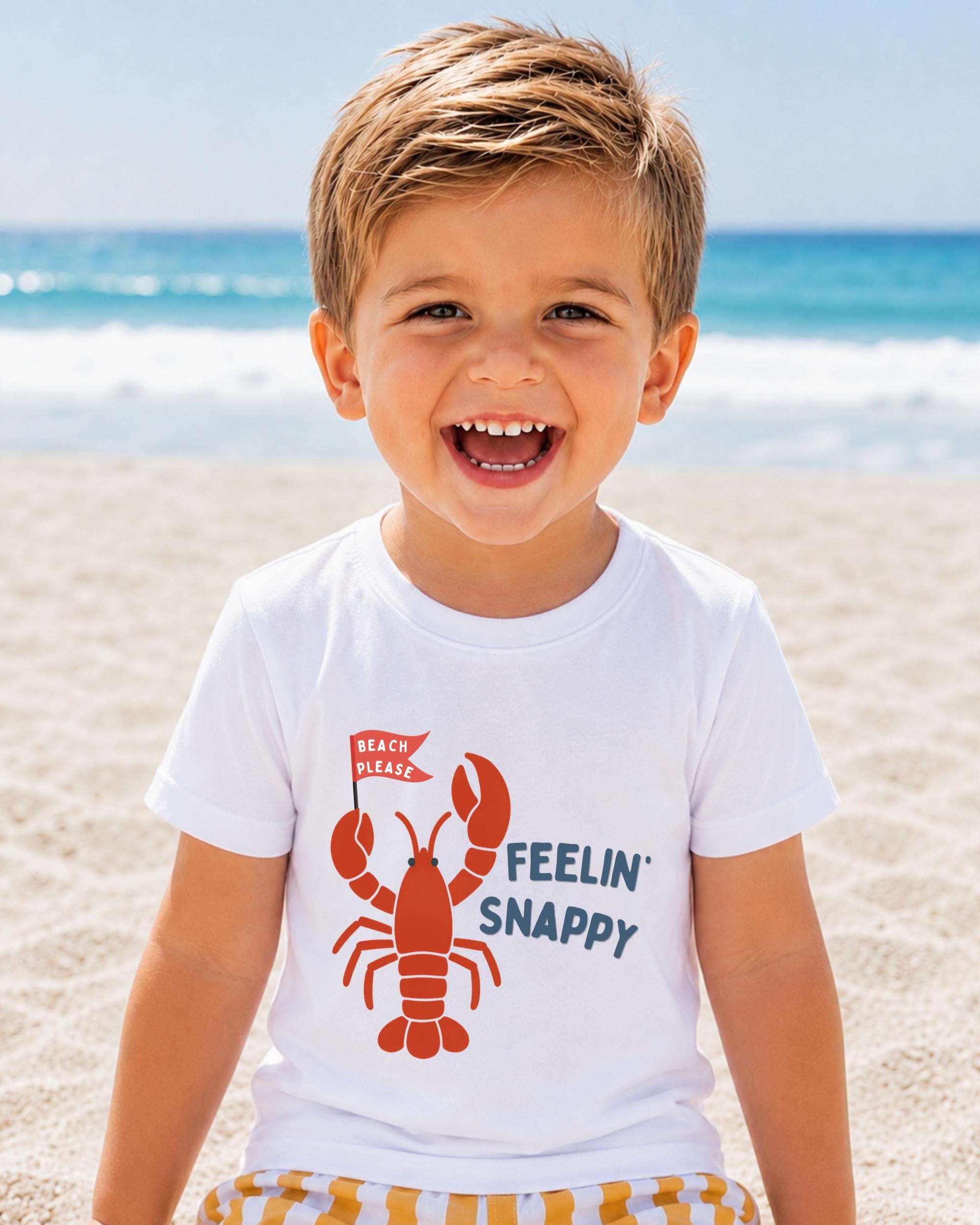 A young boy with a big smile on his face, wearing a white t-shirt with a lobster graphic and the text "Feelin' Snappy" printed on it, standing on a sandy beach with the ocean in the background.