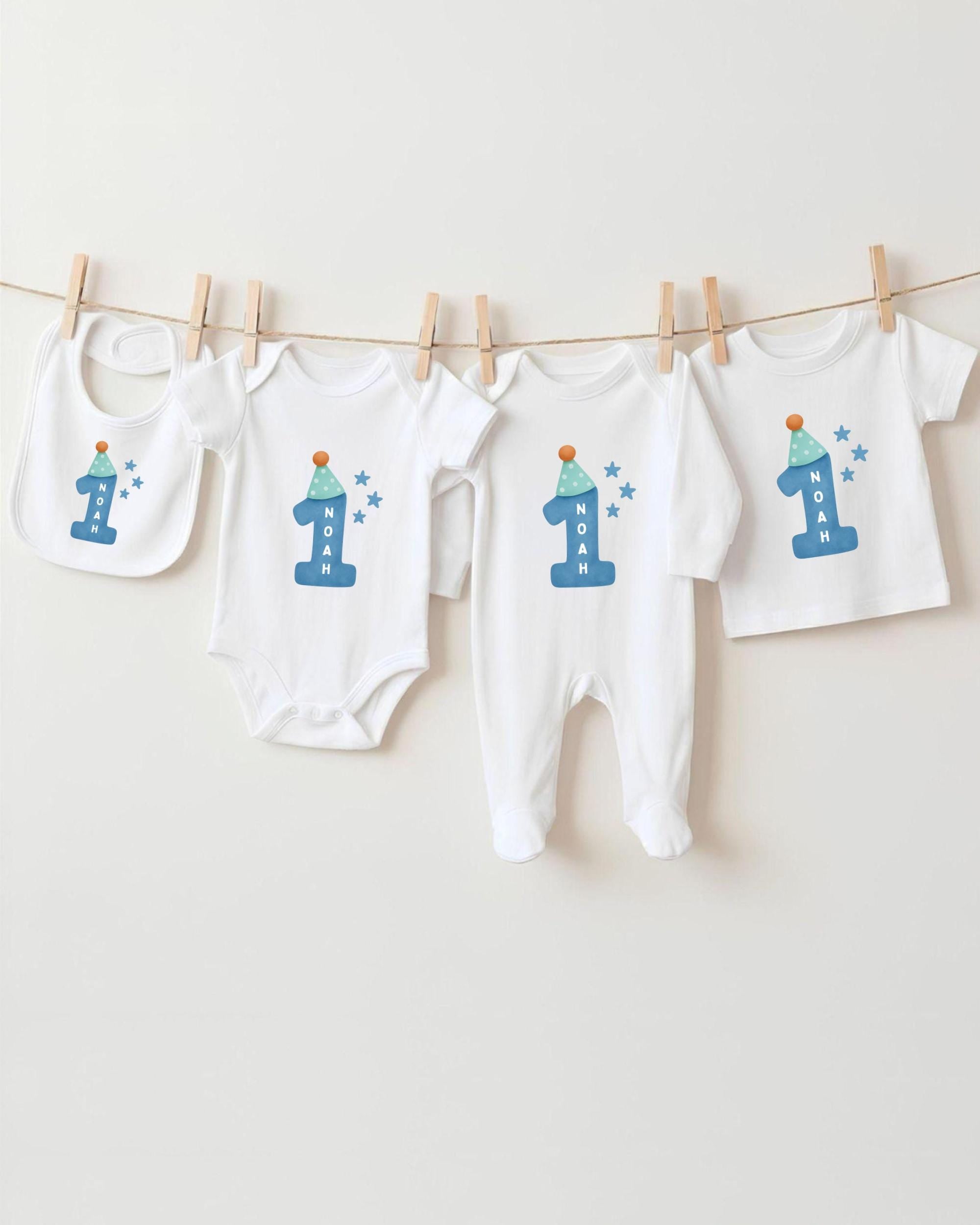 A collection of white baby clothes, including onesies and bibs, with blue and white designs and the number 1 printed on them.