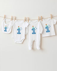 A collection of white baby clothes, including onesies and bibs, with blue and white designs and the number 1 printed on them.