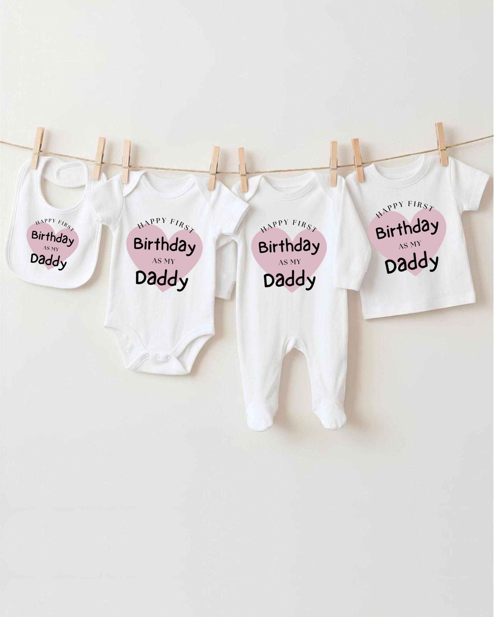 A collection of white baby clothes and bibs with the text "Happy First Birthday as my Daddy" printed on them, hanging on a clothesline.