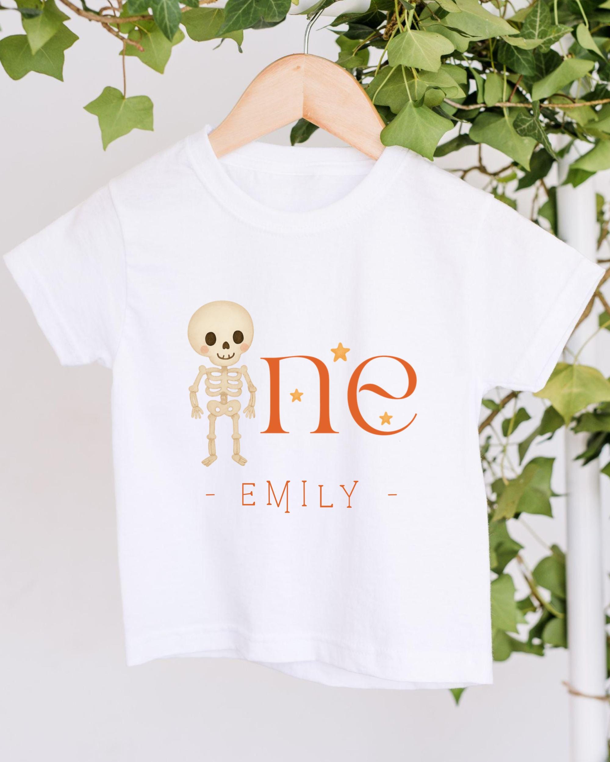 A white t-shirt with a skeleton design and the text "One - Emily" printed on it, hanging on a wooden hanger.
