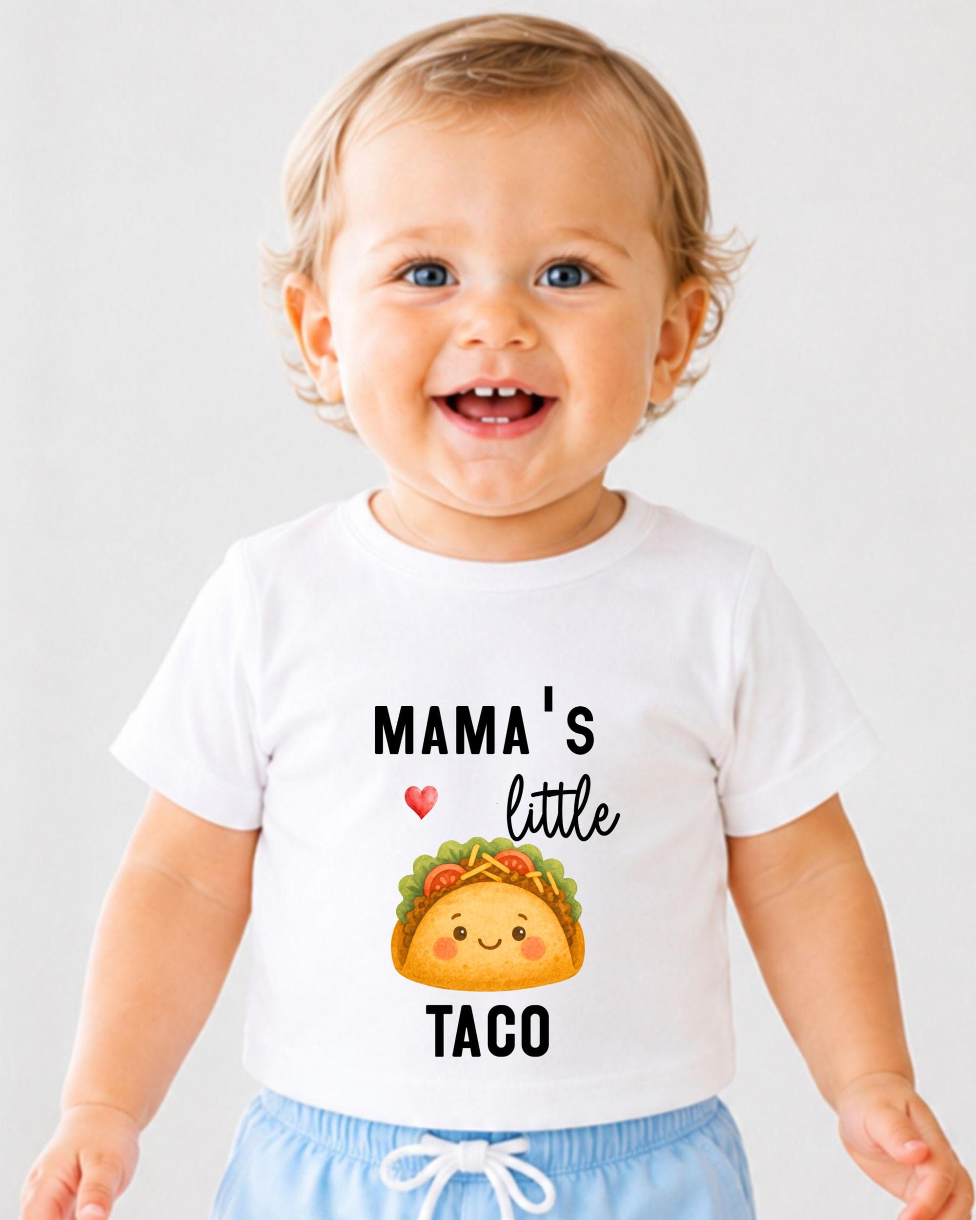 A smiling baby wearing a white t-shirt with a graphic of a taco and text that says "Mama's little taco".