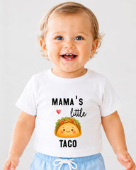A smiling baby wearing a white t-shirt with a graphic of a taco and text that says "Mama's little taco".