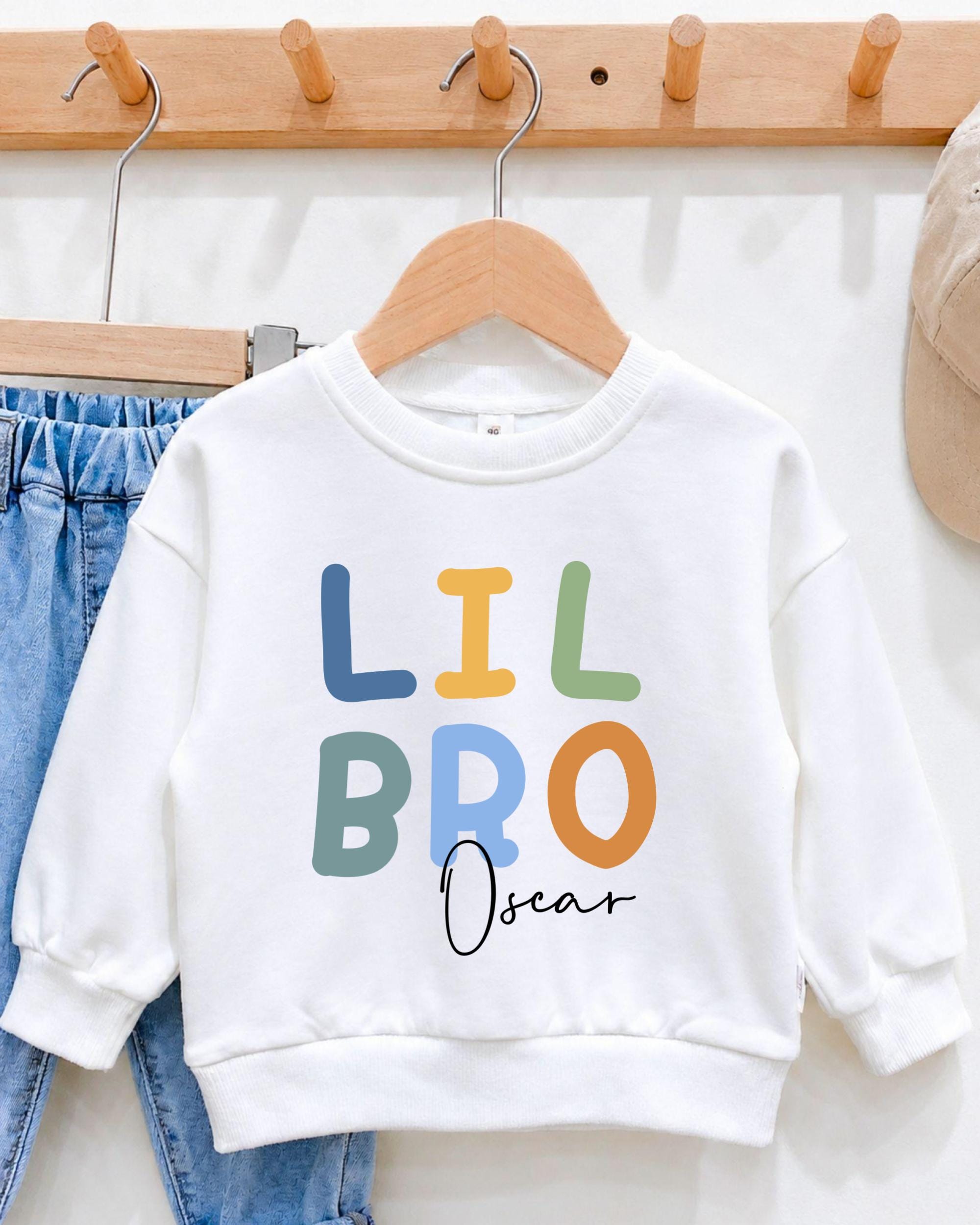 A white sweatshirt with the text "LIL BRO Oscar" printed on it, hanging on a wooden hanger.