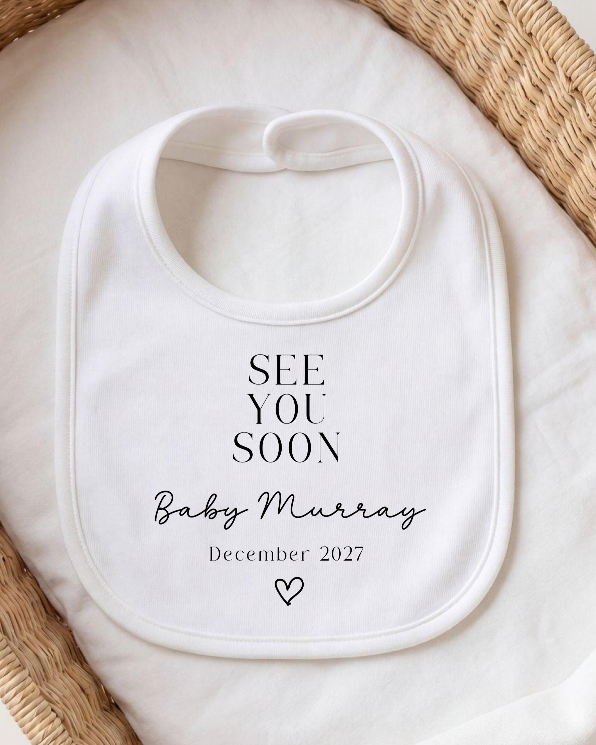 A white bib with the text "SEE YOU SOON Baby Murray December 2027" printed on it, placed on a white cloth.
