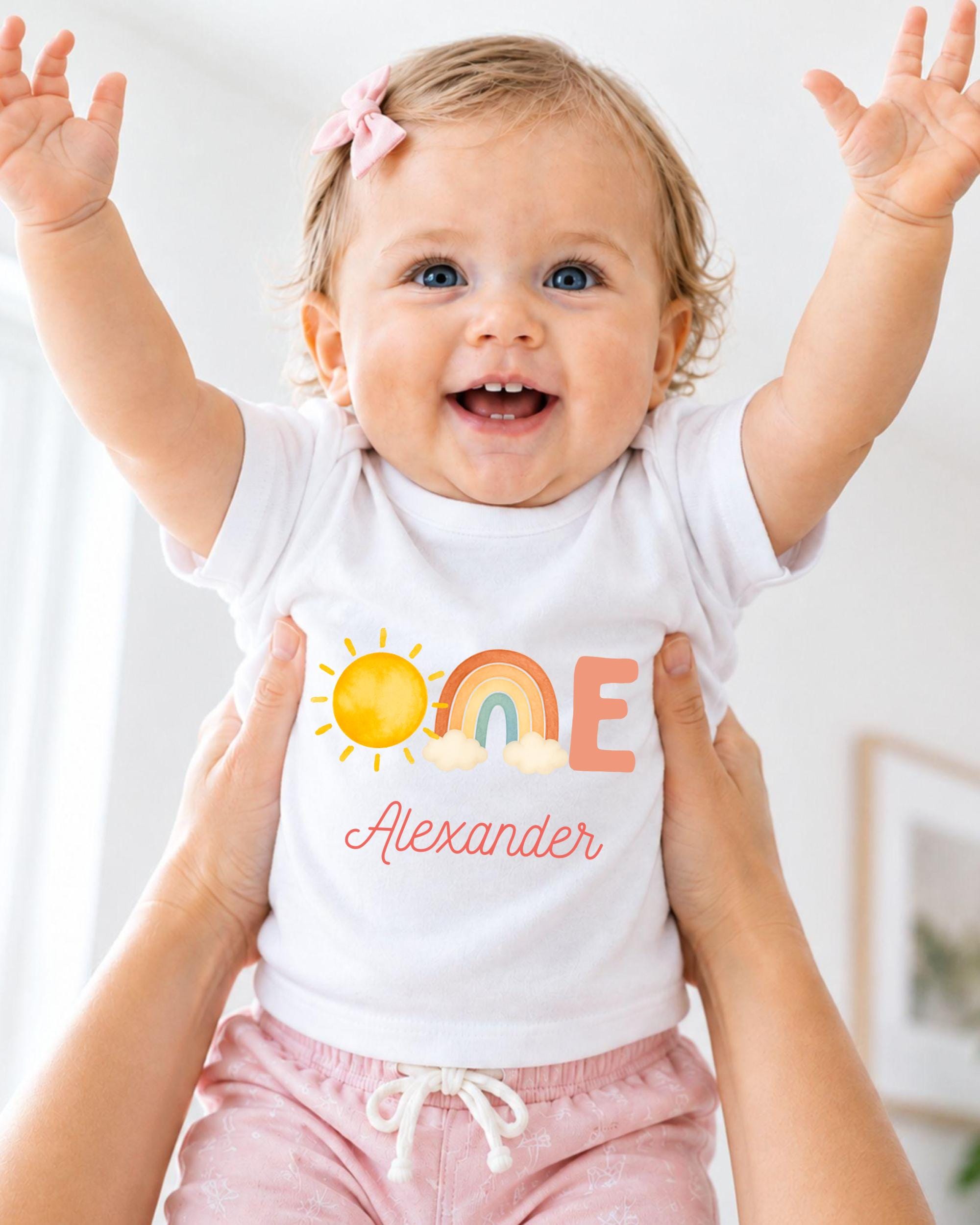 A smiling baby girl wearing a white shirt with a personalized name and a sun and rainbow design.
