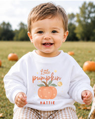 A smiling baby boy wearing a white sweatshirt with a pumpkin graphic and the text "Little pumpkin" and "HATTIE" printed on it, sitting in a field with pumpkins in the background.