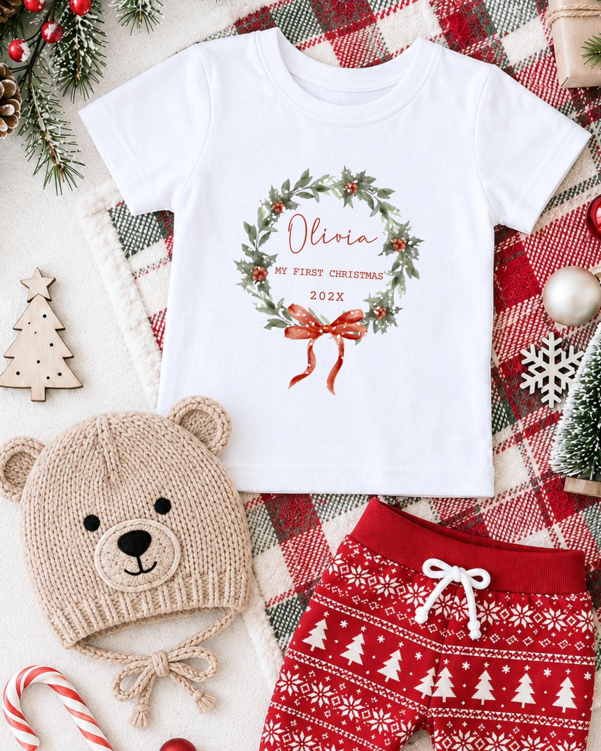 A white t-shirt with a Christmas wreath design, a beige knit hat with a bear face, red and white patterned shorts, and other holiday decorations are arranged on a plaid blanket.