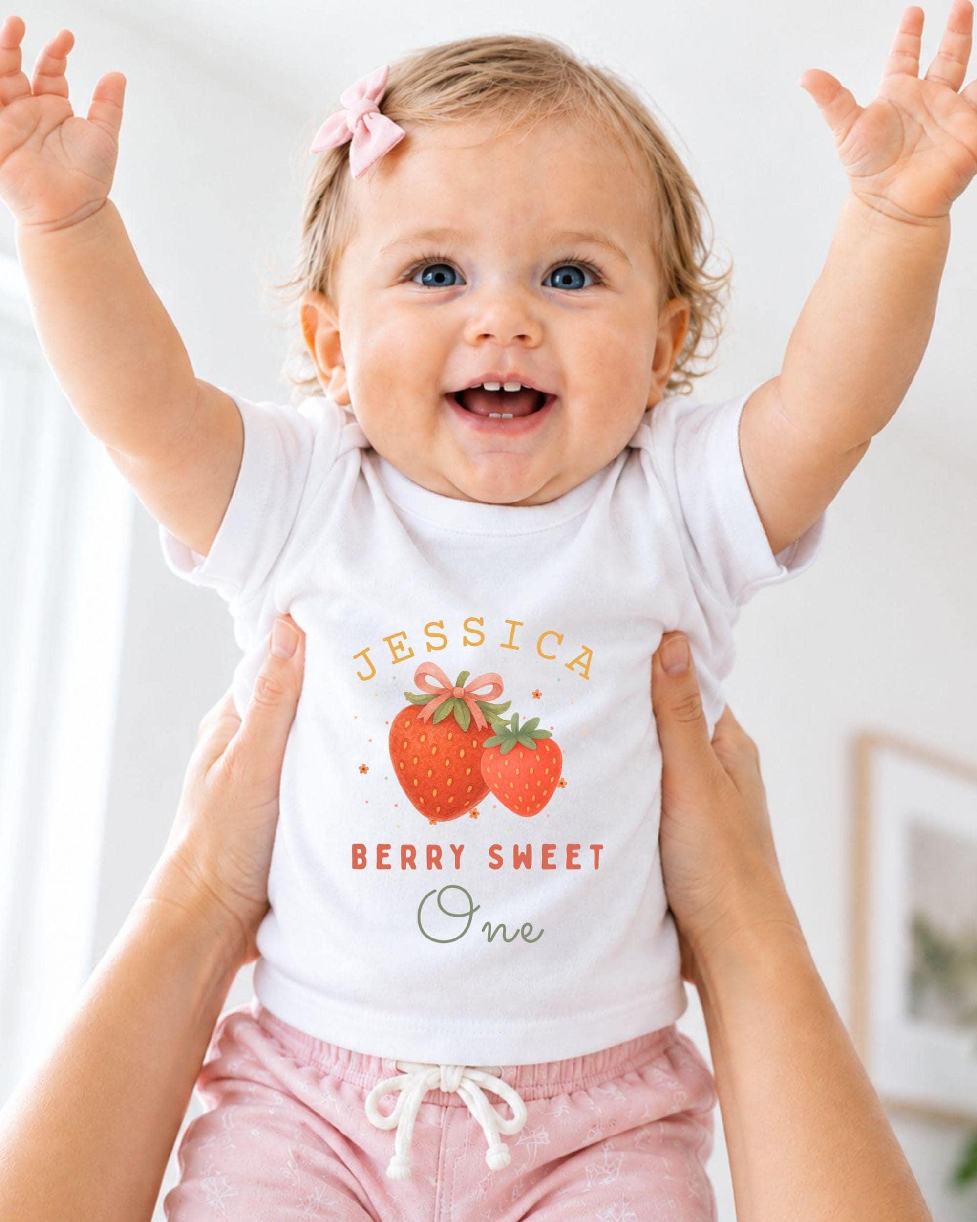 A smiling baby girl wearing a white t-shirt with a strawberry design and the name "JESSICA" printed on it, being held up by someone's arms.