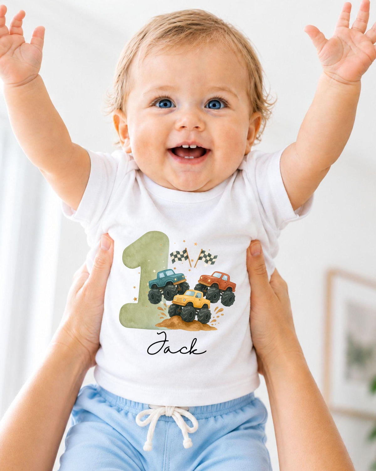 A smiling baby wearing a white t-shirt with a truck design and the name "Jack" printed on it, being held up by someone's arms.