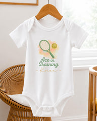 A white baby onesie with a tennis racket and ball graphic printed on it, hanging on a wooden hanger.