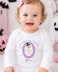 A smiling baby girl wearing a white onesie with a skeleton and the text "My first Halloween Anna" is sitting in front of Halloween decorations.