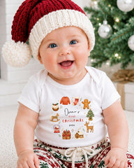 A smiling baby wearing a Santa hat and a t-shirt with a Christmas-themed design, sitting in front of a Christmas tree.