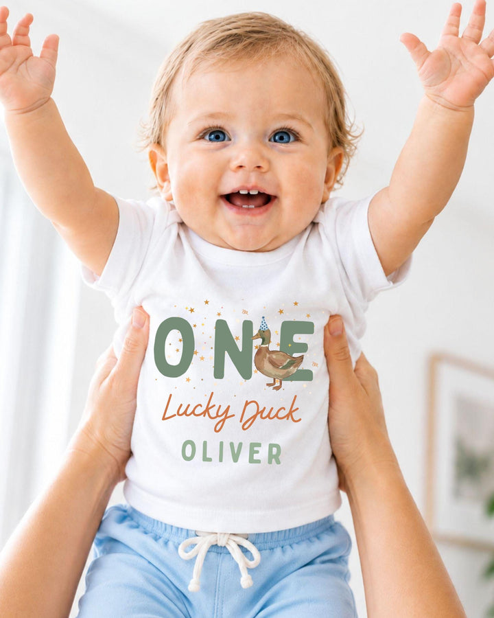 A smiling baby wearing a white shirt with the text "ONE Lucky Duck Oliver" is being held up by someone's arms.